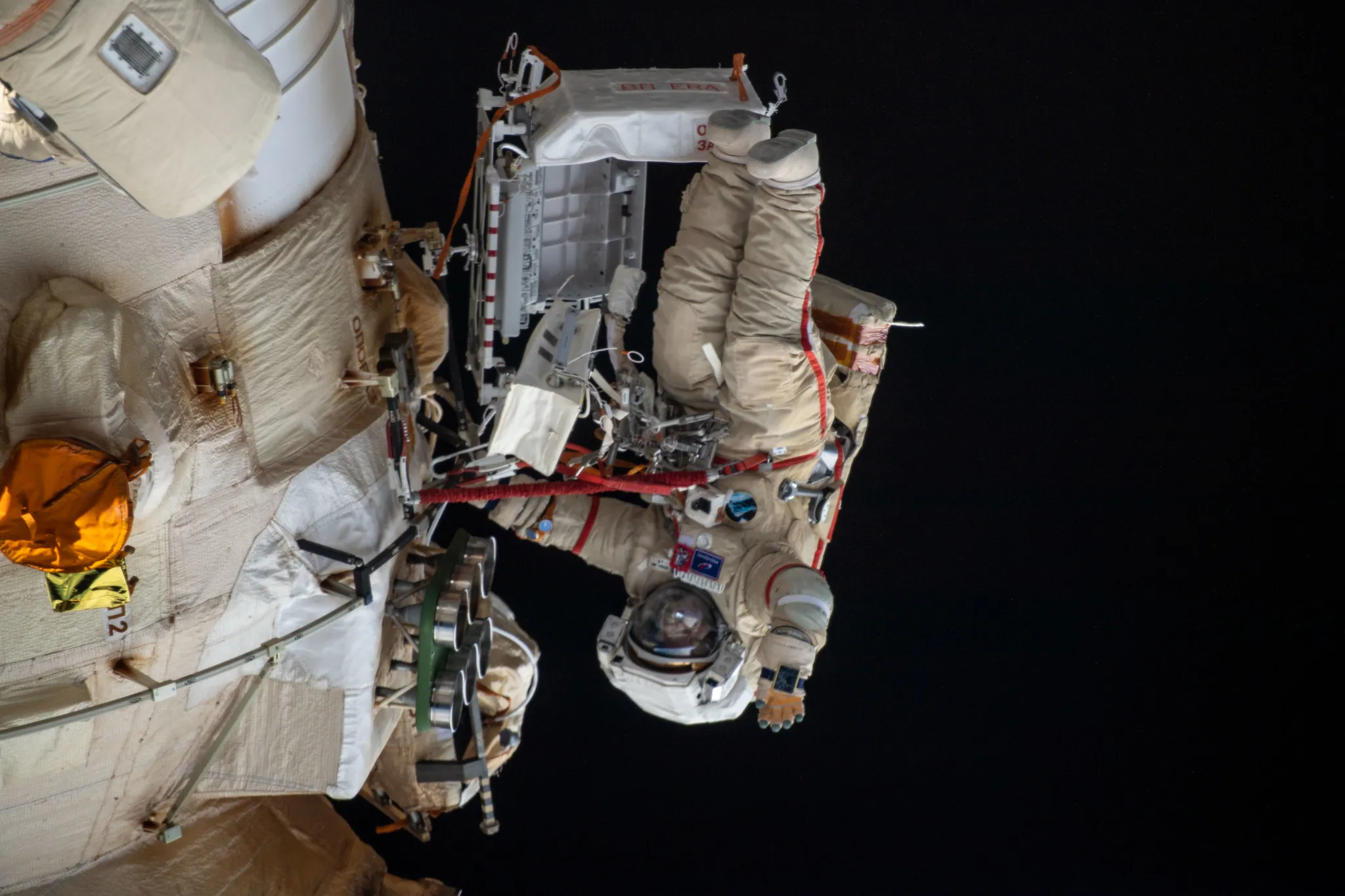 (April 18, 2022) — Cosmonaut Oleg Artemyev waves to the camera while working outside the Nauka multipurpose laboratory module during a spacewalk that lasted for six hours and 37 minutes to outfit Nauka and configure the European robotic arm on the International Space Station’s Russian segment.